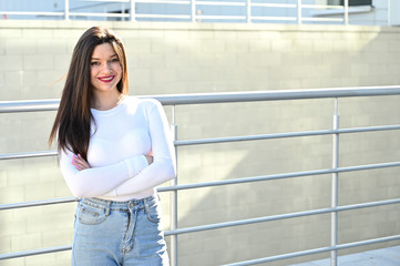 Wall Mural - Caucasian portrait of a pretty brunette girl in a white blouse and blue jeans stands near the railing with a smile against the background of a gray wall of a building on a sunny spring day.