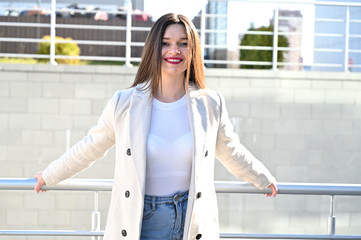 Wall Mural - Caucasian portrait of a lovely brunette girl in a light coat and blue jeans with a smile stands near the railing on a sunny spring day.