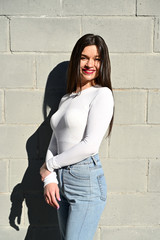 Wall Mural - Vertical Caucasian portrait of a pretty brunette girl in a white blouse and blue jeans stands near the wall of a building on a spring day in the city. Model is happy to pose.