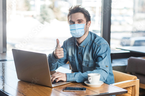 Fotomural Everything alright! Portrait of satisfied young man with surgical medical mask in blue shirt are sitting and working on laptop, thumb up, looking at camera