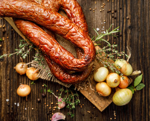 Wall Mural - Smoked sausage on a wooden rustic table with addition of fresh aromatic herbs and spices, top view. Natural product from organic farm, produced by traditional methods