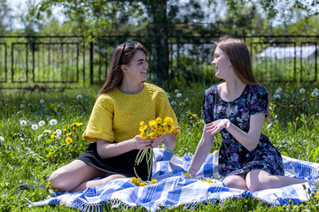 Young sisters in nature weave a wreath of dandelions. Talk and enjoy the rest. Weave flowers into the hair