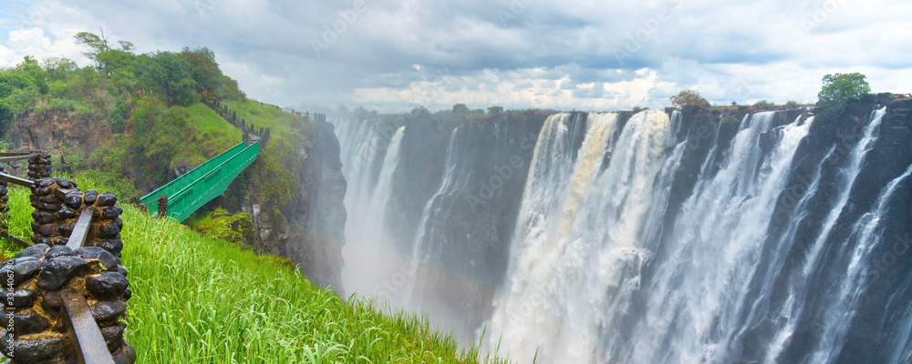 Walking way with view to the dramatic waterfall and clouds at Victoria Falls on the Zambezi River, Zimbabwe, Zambia. - obrazy, fototapety, plakaty 
