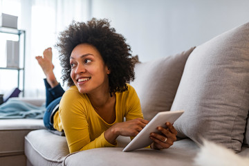 Wall Mural - Browsing a bit before the day begins. Happy young woman with tablet pc laying on sofa. Woman reading a message, e-book or information on her tablet computer