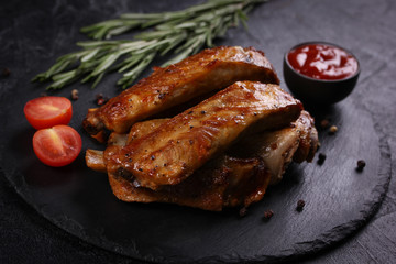 Wall Mural - Fried pork ribs with a fried crust with spices, rosemary, cherry tomatoes and ketchup in bowl on a black board on a black background. Meat on grill. Background image, copy space