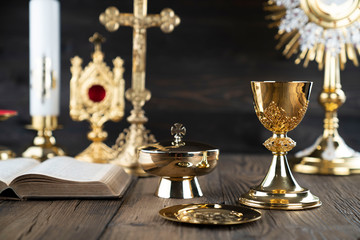 Wall Mural - Catholic religion concept. Catholic symbols composition. The Cross, monstrance,  Holy Bible and golden chalice on wooden altar and gray background. 