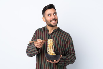 Wall Mural - Young man over isolated background holding a bowl of noodles with chopsticks and looking up