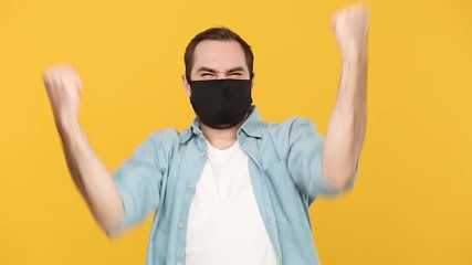 Wall Mural - Young man in sterile black face mask denim white shirt posing isolated on yellow background studio. Epidemic pandemic coronavirus 2019-ncov sars covid-19 virus concept. Happy guy doing winner gesture.