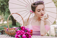 Romantic Young Pretty Woman In Pink Retro Dress And Pin Up Hairstyle Sitting At Table With Basket Of Peonies Flowers And Holding Umbrella In Hand. Tender Vintage Girl On Picnic On Summer Garden