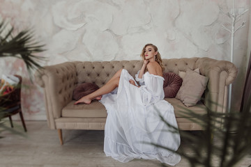 Fashion bride in a fashionable white dress with her hair and long earrings sitting on a modern sofa