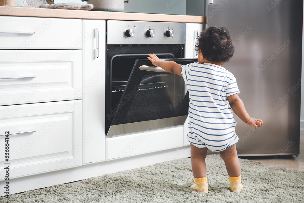 Little African-American baby near stove in kitchen. Child in danger - obrazy, fototapety, plakaty 