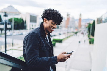 Happy Latino male tourist using public internet for sharing journey expressions to social website for travellers, excited man with cellphone device chatting via online application for communication