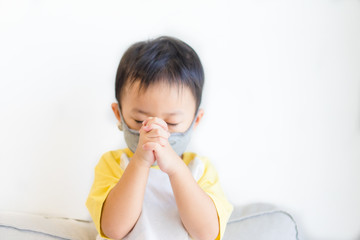 Little Chinese toddler boy wearing face mask for Covid-19 coronavirus.Prayer kid boy worship to GOD at home in sunday service.Church online.Coronavirus pandemic.Hand praying for thank GOD.Copy space.