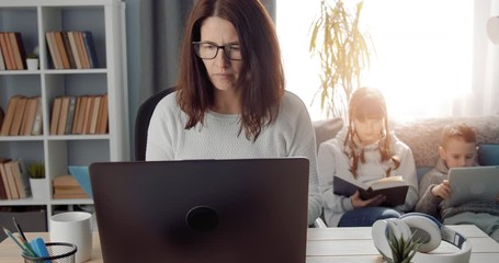 Wall Mural - Mother in eyeglasses and domestic clothing working remotely on laptop while taking care of her two children at home. Concept of coronavirus and quarantine