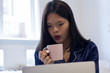 © Tripodland - Young asian smiling woman working on laptop while holding a pink mug. Happy morning