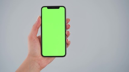 Female hands holding a smartphone with a green screen on a gray background. Chroma key. Copy Space