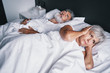 © luengo_ua - Senior couple in the bed. Pretty aged woman is angy with her sleeping husband covering ears with hands.