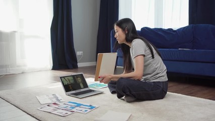 Wall Mural - Tilt up side view of Asian woman sitting cross-legged on floor at home, having video call with colleagues on laptop and sharing ideas showing him sketches in notepad and printed interface templates