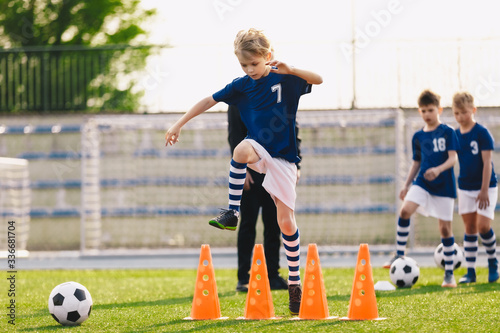 Boy jumping over soccer cones on football training field. Kids on sports training with junior coach. Children on physical education class. Boys imporoving soccer skills