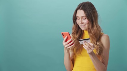 Poster - Happy attractive girl holding credit card and shopping online through smartphone isolated on blue background