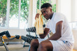 © twinsterphoto - African American sportsman sitting on bench and wiping sweat with towel. Black athlete resting during fitness workout in contemporary gym.