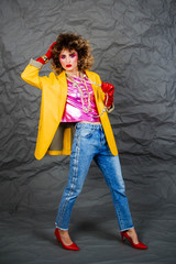 Wall Mural - Girl in a yellow jacket and blue jeans with an afro hairstyle. Fashion of the eighties, disco era. Studio photo on gray background.