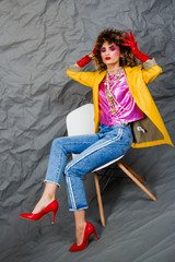 Wall Mural - A girl in a yellow jacket and blue jeans with an afro hairstyle sits on a chair. Fashion eighties, the era of disco. Studio photo on a gray background.
