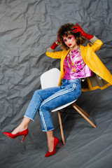 Wall Mural - A girl in a yellow jacket and blue jeans with an afro hairstyle sits on a chair. Fashion eighties, the era of disco. Studio photo on a gray background.
