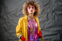 Portrait Of A Girl In A Yellow Jacket And Blue Jeans With Afro Hair Of The Eighties, Disco Era. Photo In Studio On A Gray Background.