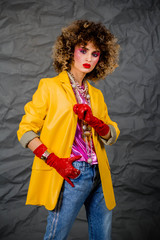 Wall Mural - Portrait of a girl in a yellow jacket and blue jeans with afro hair of the eighties, disco era. Photo in studio on a gray background.