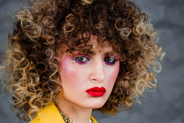 Portrait of a girl in a yellow jacket and blue jeans with afro hair of the eighties, disco era. Photo in studio on a gray background.