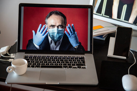 Laptop con dentro un immagine di uomo in giacca con guanti in lattine e mascherina protettrice , contestualizzato in una postazione di lavoro con altri elementi quali monitor 