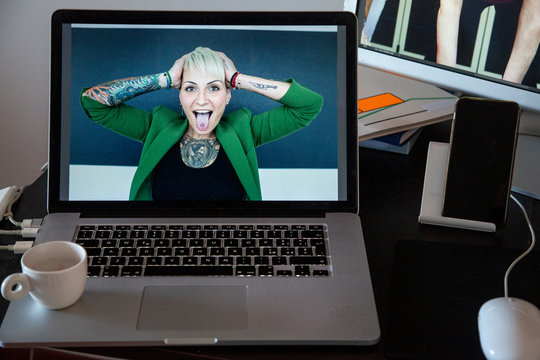 Laptop con dentro un immagine di ragazza tatuata che si tiene la testa urlando su sfondo scuro , contestualizzato in una postazione di lavoro con altri elementi quali monitor 