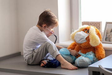 child plays a doctor. The boy listens to the breath of a toy to a large dog with a stethoscope. A toy in a medical bandage and shoe covers on legs.