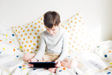 School Boy On Bed At Home With Digital Pad Tablet In Hand, Doing Homework. Distance Learning Online Education. Quarantine. Game. Boy Plays With Smartphone