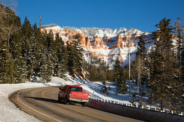 Red truck driving up winding canyon road toward desert towers of red sandstone.