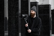 © Volodymyr - Bearded Professional videographer in black hoodie holding professional camera on 3-axis gimbal stabilizer. Filmmaker making a great video with a professional cinema camera. Cinematographer.