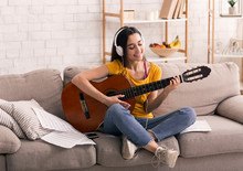 Stay Home Leisure Activities. Happy Girl With Headphones Playing Guitar On Sofa In Living Room