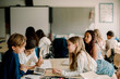 © Maskot - Female student leaning over table while friends sitting in classroom