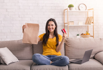 Wall Mural - Online shopping. Young smiling girl with laptop, credit card and paper bag sitting on sofa at home