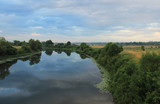 Fototapeta Na ścianę - A wide river at a bend surrounded by trees and bushes. Blue clouds are reflected in the surface of the river.