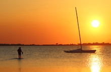 Silhouette Of Person In Backlight Walking Into The Sea At Low Tide Towards A Sailboat At Sunset