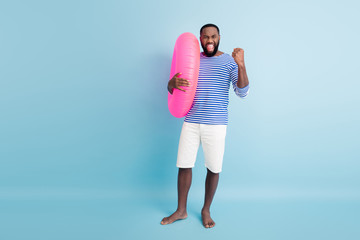 Wall Mural - Full length photo ecstatic afro american man celebrate holiday lottery victory raise fists scream yes hold pink float circle wear white shorts nautical vest isolated blue color background