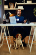 © chika_milan - Cheerful smiling handsome Caucasian man sitting in his home office with dog sitting under the table.