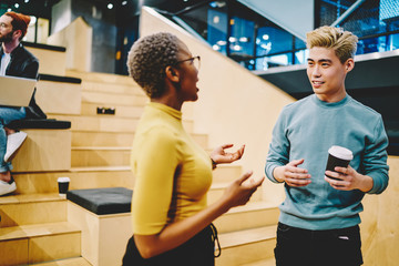 Multiracial male and female young employees having conversation during coffee break enjoying friendly atmosphere, chinese man student in smart casual wear listening to his dark skinned colleague talk