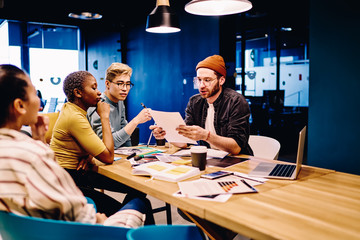 Young caucasian male leader of working group reading documents and analyzing data for project organization, skilled diverse team of designers planning brainstorming session sitting at table in office