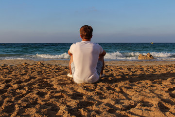 a young man of athletic build in a white t-shirt sits on the beach or ocean on golden sand and looks