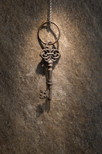 Old Vintage Door Or Chest Key Hanging On A Chain Against The Background Of A Wall In A Ray Of Light, Concept Of Mystery, Treasure, Mystery