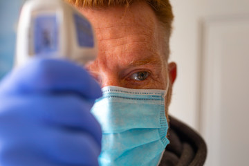 Man in a face mask and gloves checking body temperature with infrared forehead thermometer.