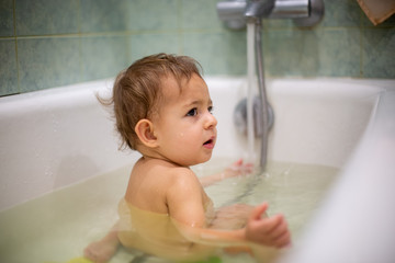 Wall Mural - A cute Caucasian baby takes a bath sits in water with his back to the camera and turned his head toward the parent. close-up, soft focus, blur background